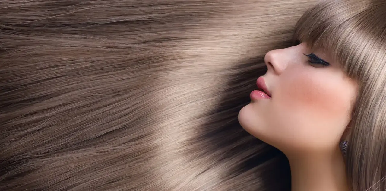 Kobieta z subtelnym makijażem i fryzurą w odcieniu beżowy blond sombre, otoczona gęstymi, lśniącymi włosami.