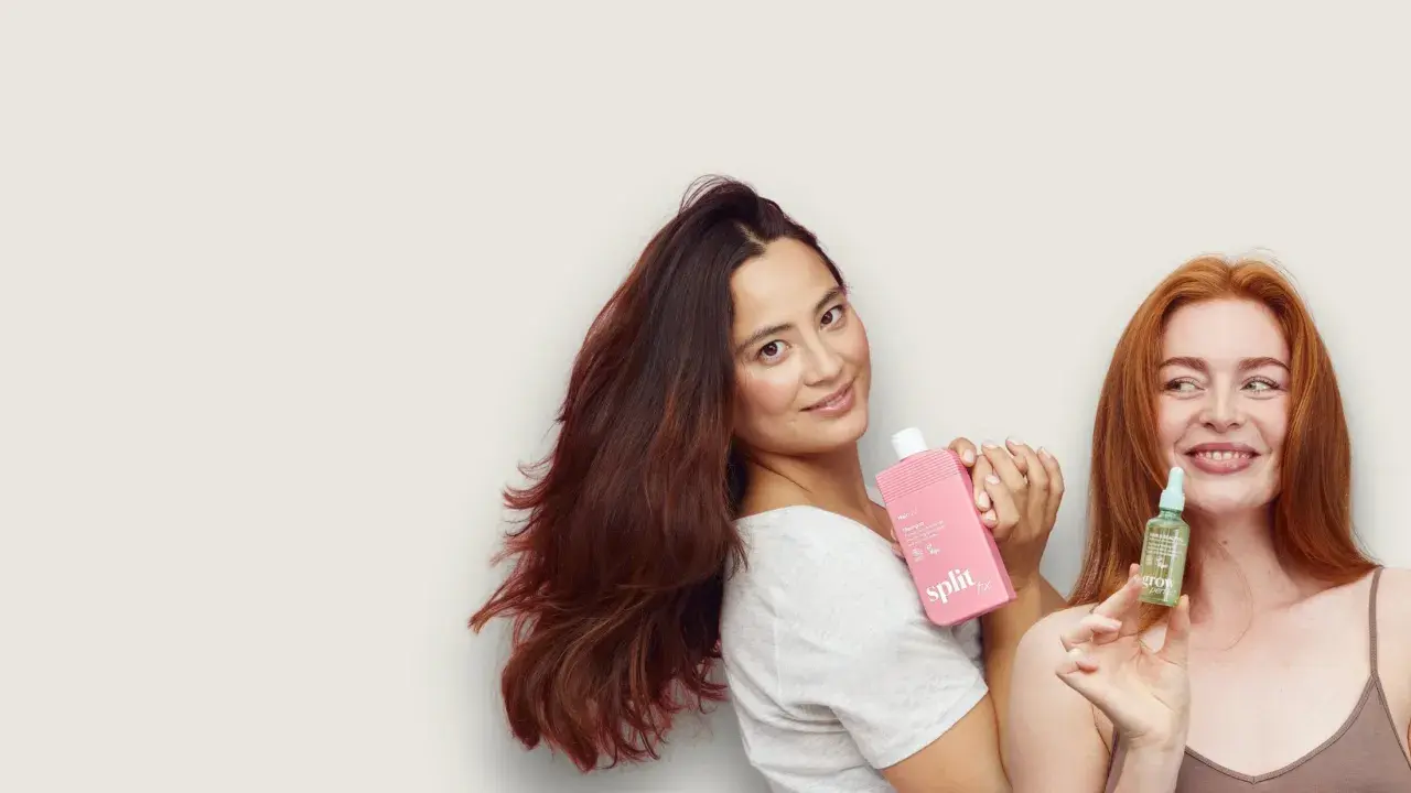 Kobiety prezentują kosmetyki do włosów. Jedna trzyma różową butelkę, druga małą buteleczkę z serum, które zawiera olejek jojoba właściwości odżywcze.