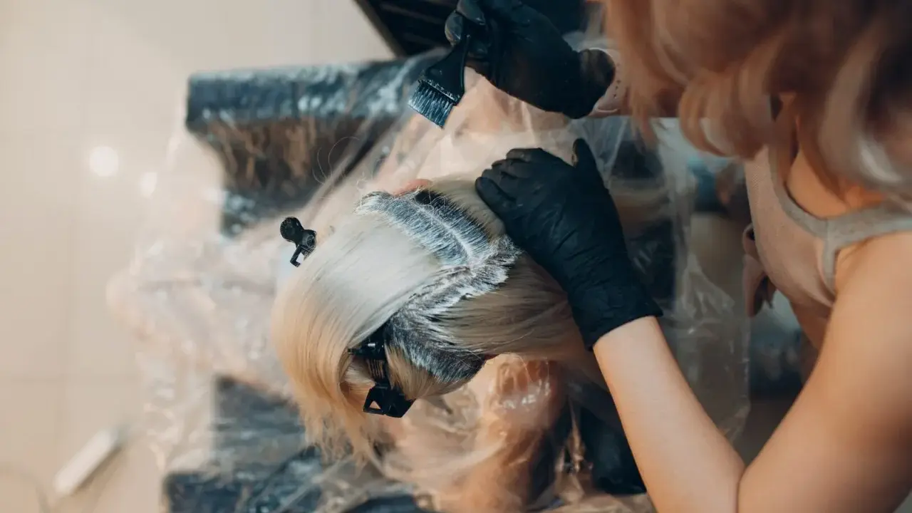 Fryzjerka w czarnych rękawiczkach nakłada farbę na blond włosy. Kluczowe jest, ile trzymać farbę na włosach, by uzyskać idealny efekt.