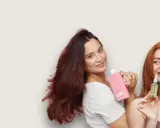 Kobiety prezentują kosmetyki do włosów. Jedna trzyma różową butelkę, druga małą buteleczkę z serum, które zawiera olejek jojoba właściwości odżywcze.