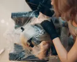 Fryzjerka w czarnych rękawiczkach nakłada farbę na blond włosy. Kluczowe jest, ile trzymać farbę na włosach, by uzyskać idealny efekt.