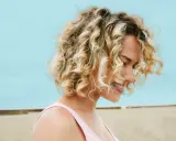 Uśmiechnięta blondynka z burzą loków na plaży. Zastanawiasz się, ile trwa zrobienie trwałej? To zależy od wielu czynników!