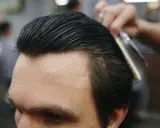 Fryzjer układa włosy mężczyzny na gładki slick back grzebieniem i nożyczkami.