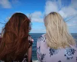 Dwie kobiety z włosami: jedna z ciemnymi, druga po farbowaniu ciemnych włosów na jaśniejsze, blond. Wiatr rozwiewa ich włosy nad morzem.