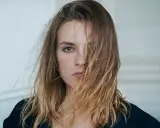 Kobieta z burzą blond włosów, które zasłaniają jej twarz. Włosy wyglądają na lekko potargane, jakby wiatr je rozwiewał, tworząc efekt strączkowania włosów.