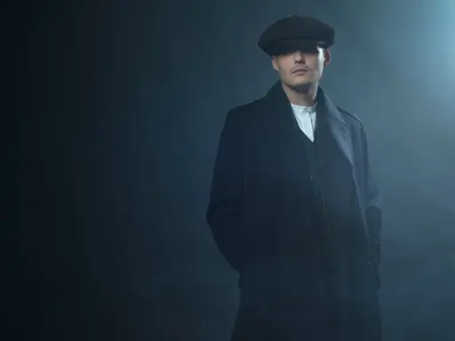 Młody mężczyzna w czapce i płaszczu, z charakterystyczną fryzurą w stylu Peaky Blinders, stoi w zadymionym mroku.
