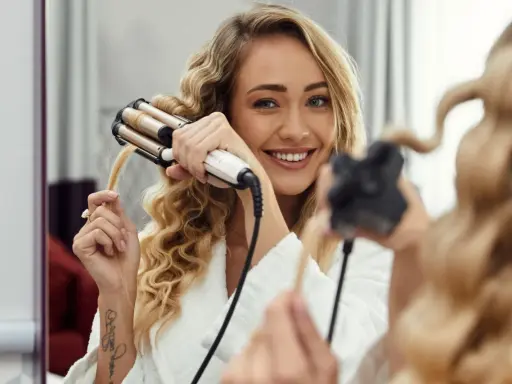 Uśmiechnięta blondynka w szlafroku tworzy piękne fale na włosach za pomocą karbownicy.