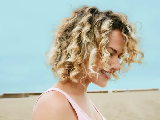 Uśmiechnięta blondynka z burzą loków na plaży. Zastanawiasz się, ile trwa zrobienie trwałej? To zależy od wielu czynników!