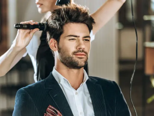 Mężczyzna w eleganckim garniturze czeka na stylizację fryzury męskiej. Fryzjerka suszy jego włosy suszarką.