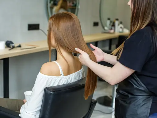 Fryzjerka wykonuje degażowanie włosów krok po kroku. Klientka siedzi w fotelu, a fryzjerka delikatnie rozdziela jej długie, rude włosy.