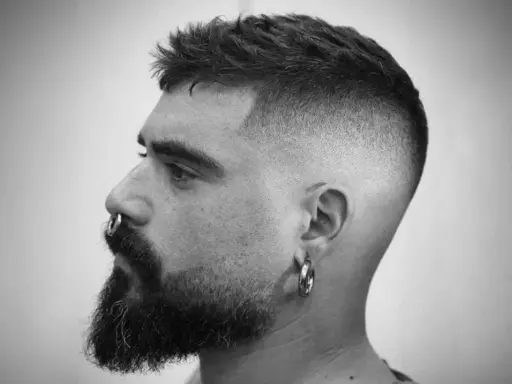 Mężczyzna z brodą i kolczykiem w nosie, z modnym, dłuższym buzz cut i cieniowanymi bokami.