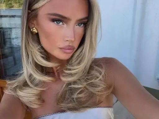 Kobieta z blond włosami, stylizowanymi na fale, z subtelnym makijażem i złotymi kolczykami. Jej fryzura przypomina buzz cut, ale jest dłuższa i bardziej objętościowa.